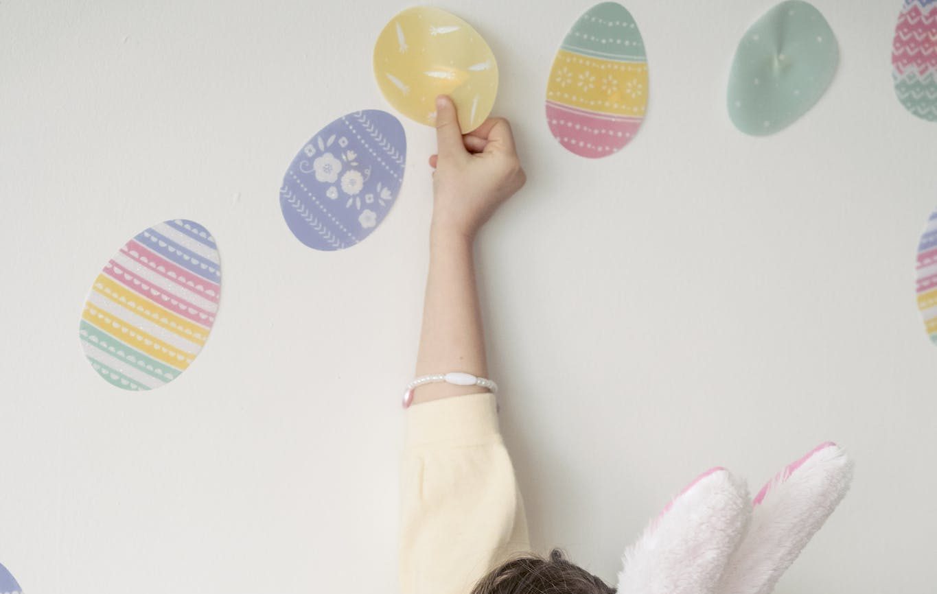 Side view content little girl wearing bunny ears applying colorful Easter stickers on white wall.