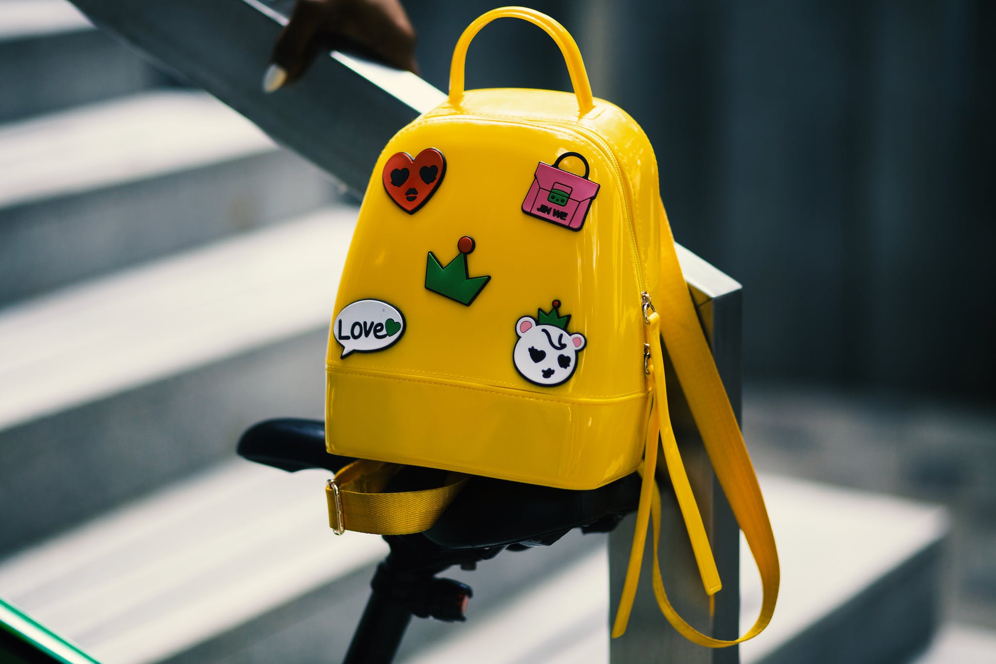 Yellow Backpack With Five Assorted Stickers on Grey Metal Stairway Rail.